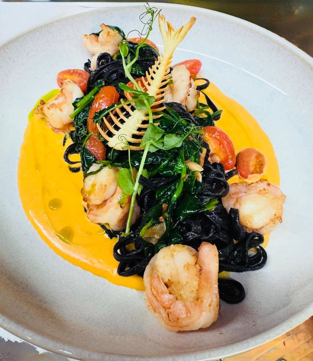 Apfelbaum – Restaurant – Dieses Gericht sind Tintenfisch-Tagliolini, serviert mit Garnelen und Kirschtomaten auf einer leuchtend gelben Sauce, garniert mit einem markanten Fischskelett.
